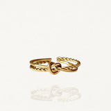 Bague fine ouverte en acier inoxydable doré avec motif nœud central et anneau effet torsadé sur fond blanc.