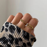 Bague nœud Alberta portée en accumulation (stacking) avec d'autres bagues fines dorées sur une main avec pull léopard.