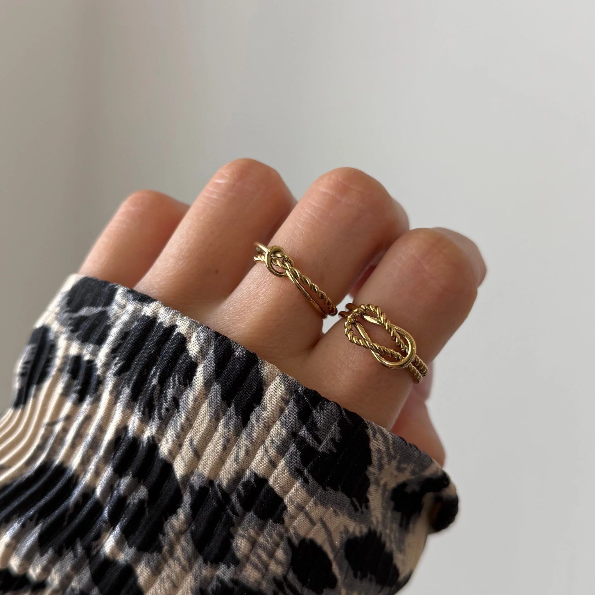 Bague nœud Alberta portée en accumulation (stacking) avec d'autres bagues fines dorées sur une main avec pull léopard.