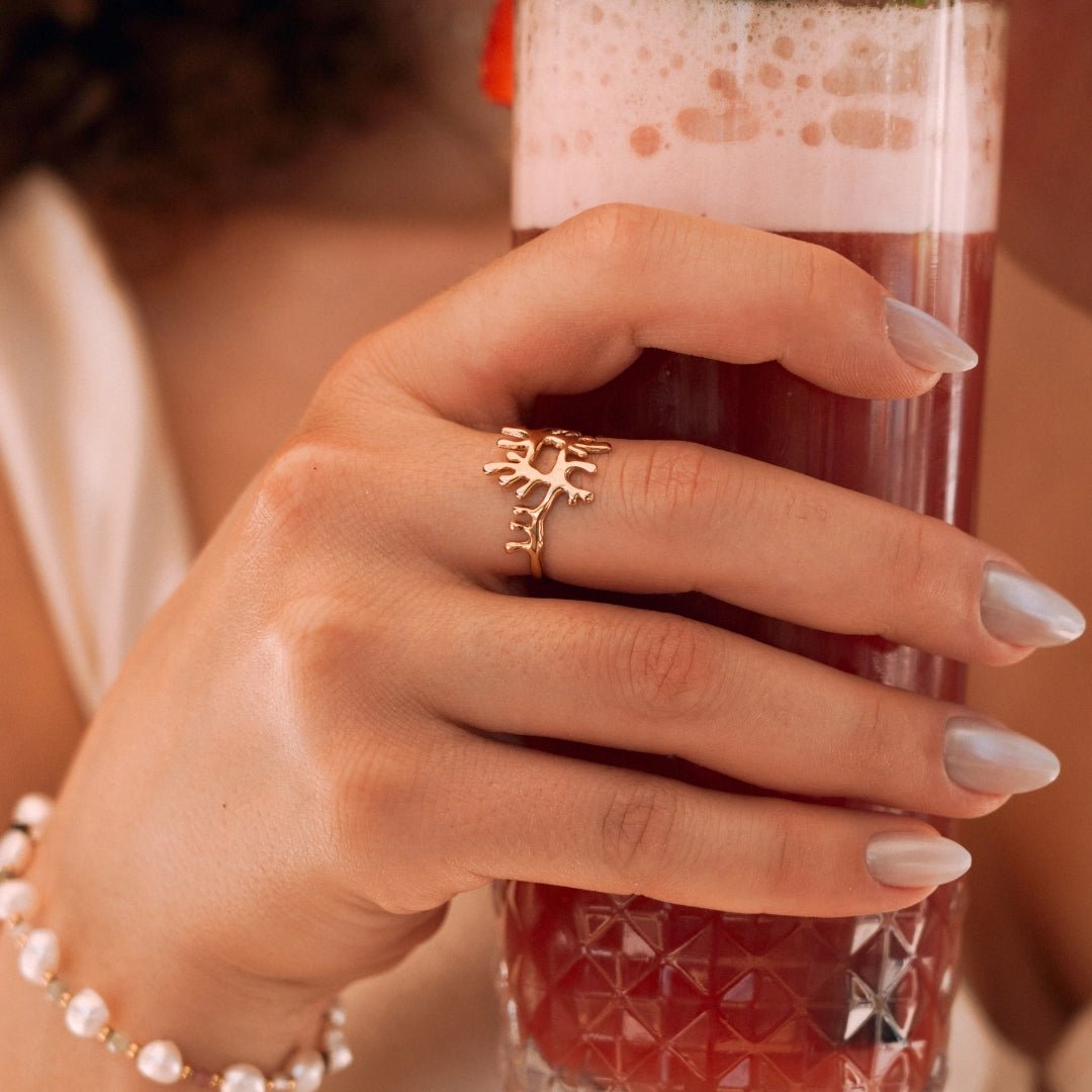 Bague en acier inoxydable doré design branches de corail portée par une femme tenant un verre de jus rouge.