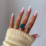Bague Cataleya portée en accumulation (stacking) avec d'autres bagues dorées et pierres naturelles vertes (style malachite ou aventurine).