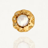 Gros plan sur une bague dorée martelée, de forme organique et irrégulière, ornée d'un cabochon central rond en résine blanche irisée effet nacre, sur fond clair.