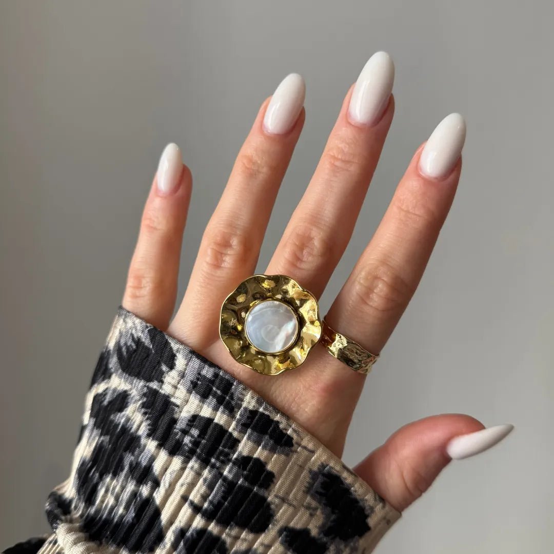 Large bague dorée martelée nacre blanche sur majeur, bague dorée texturée sur annulaire, main aux ongles blancs longs.