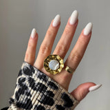 Large bague dorée martelée nacre blanche sur majeur, bague dorée texturée sur annulaire, main aux ongles blancs longs.