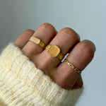 Bague Christina portée en accumulation (stacking) avec une chevalière ovale et d'autres bagues dorées.