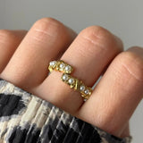 Bague ouverte dorée avec 8 perles blanches, alignées par paires dans des motifs rectangulaires ornés de croisillons, portée sur un doigt.