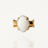 Bague dorée sertie d'une large pierre blanche ovale aspect givré ou craquelé avec fixation à griffes sur fond blanc.