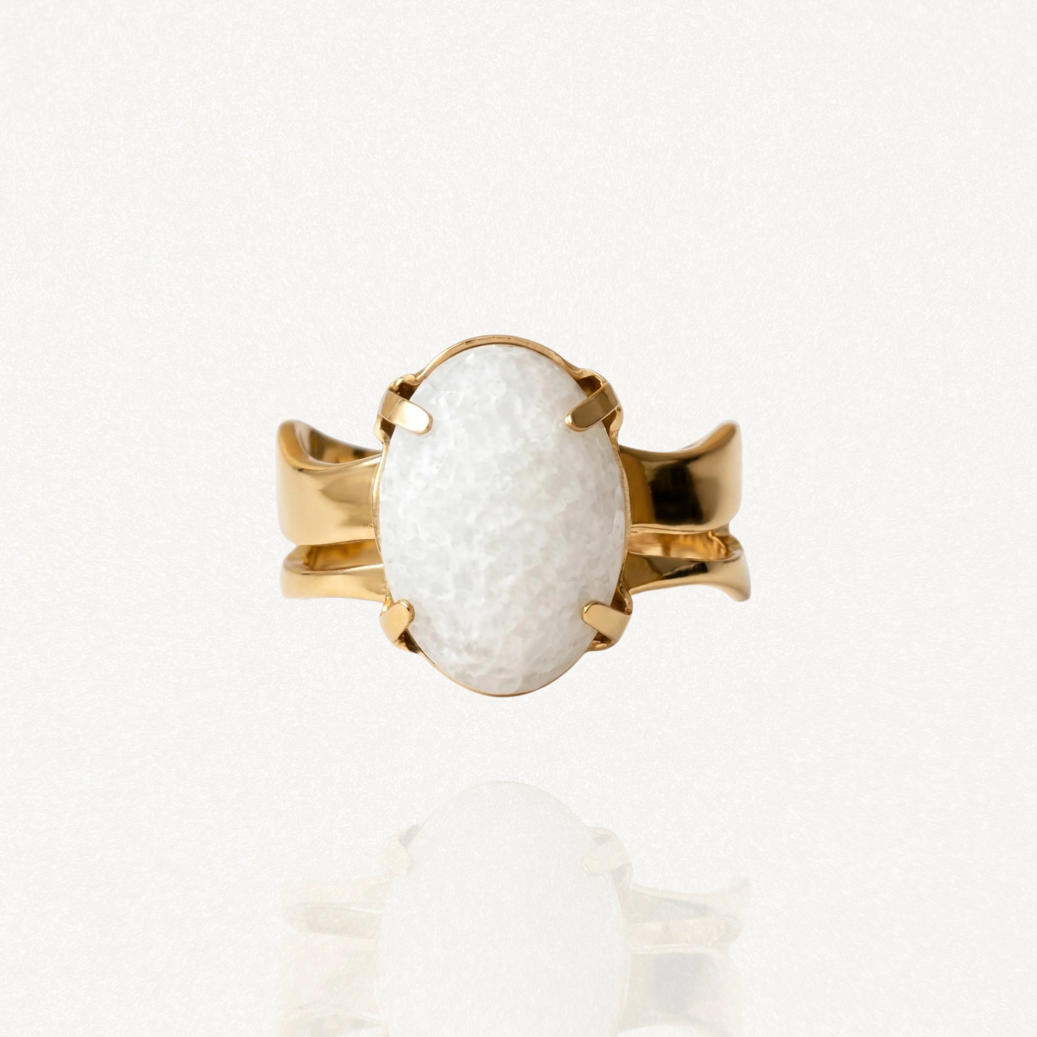 Bague dorée sertie d'une large pierre blanche ovale aspect givré ou craquelé avec fixation à griffes sur fond blanc.