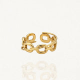 Une bague ouverte dorée en acier inoxydable avec un motif de maillons de chaîne entrelacés et une bande texturée, posée sur une surface blanche réfléchissante.