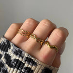 Main sur vêtement léopard, portant trois fines bagues dorées ondulées, une bague dorée maillons infini et une bague forme cornetto.