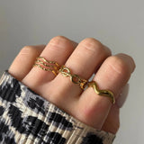 Main sur vêtement léopard, portant trois fines bagues dorées ondulées, une bague dorée maillons infini et une bague forme cornetto.