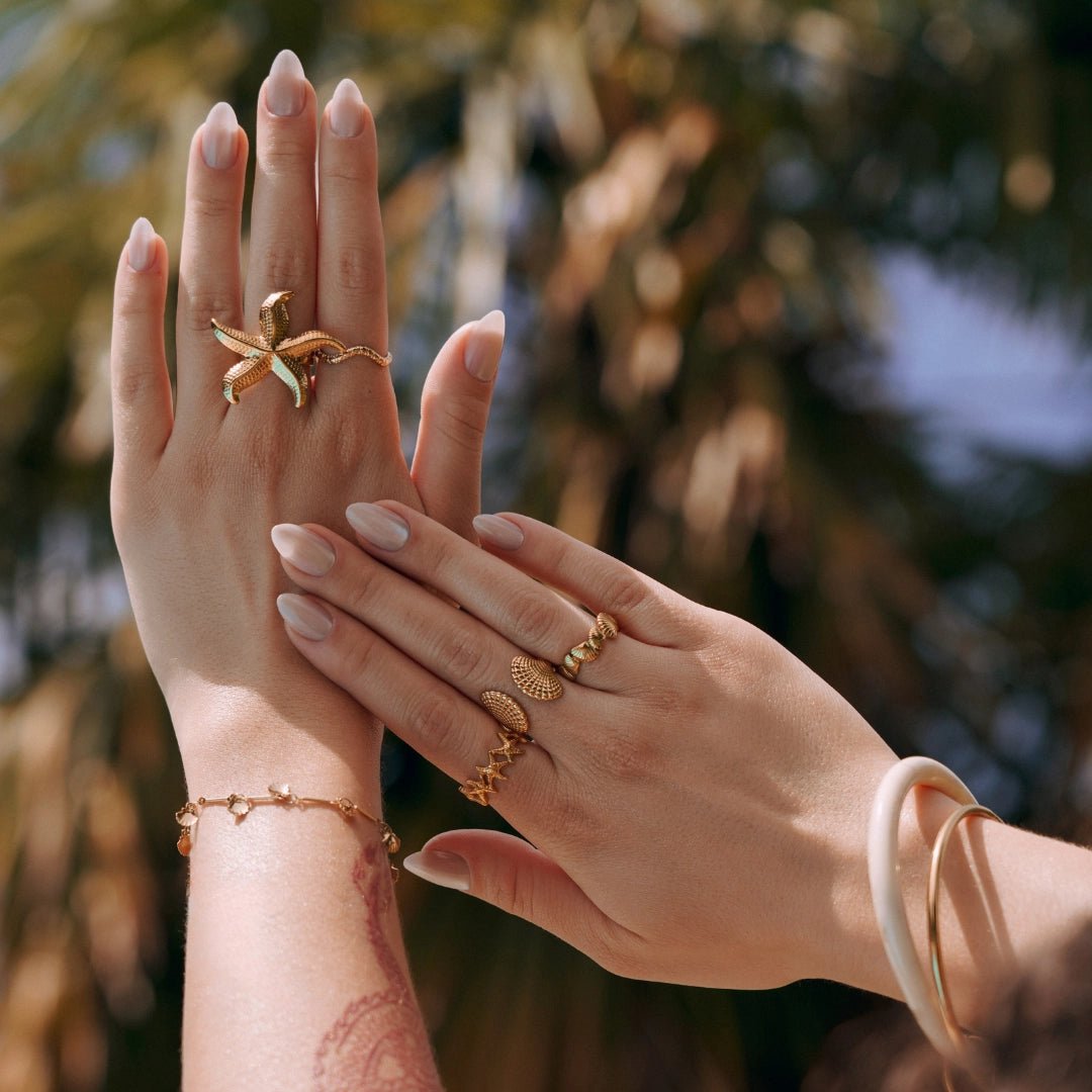 Bagues dorées en acier inoxydable sur mains de femme: bague étoile de mer, bague coquillage, et bagues fines. Style été.