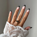 Gros plan bague Inès à pierres noires style onyx portée au doigt, contraste chic avec une manche en dentelle blanche.