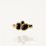 Bague fine en acier inoxydable doré ornée d'une composition de trois petites pierres noires (formes géométriques) sur fond blanc.