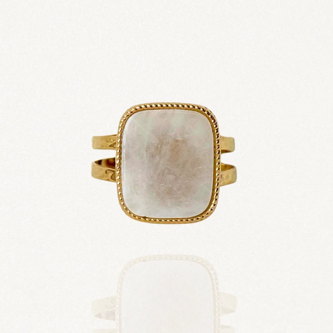Bague large en acier inoxydable doré ornée d'une pierre carrée blanche effet nacre et bordure perlée sur fond blanc.