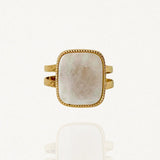 Bague large en acier inoxydable doré ornée d'une pierre carrée blanche effet nacre et bordure perlée sur fond blanc.