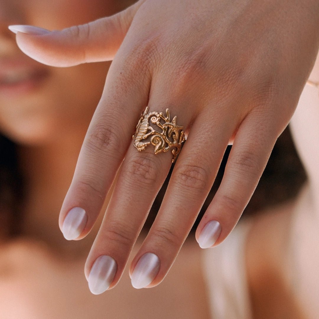 Bague en acier inoxydable doré, motif marin détaillé : hippocampe, corail, étoile de mer, portée sur une main.