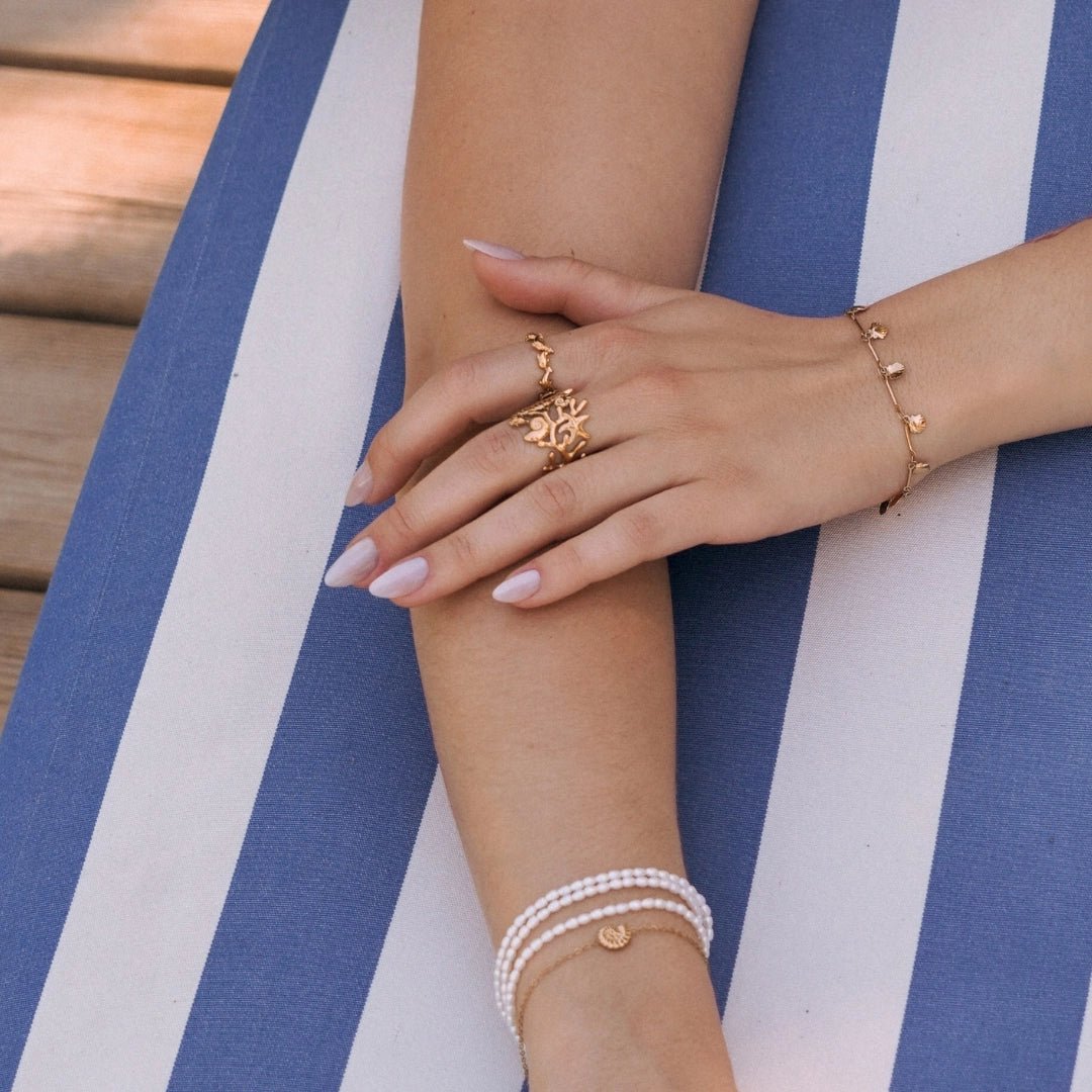 Bague en acier inoxydable doré, design corail ajouré, portée par une femme sur fond rayé bleu et blanc.