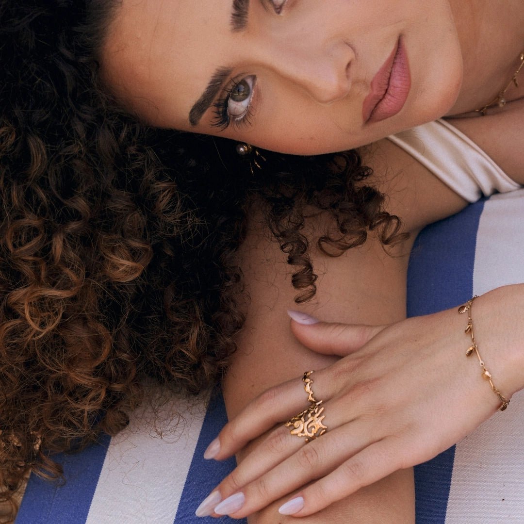 Bague en acier inoxydable dorée à motif soleil portée par une femme, sur fond rayé bleu et blanc.