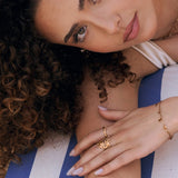 Bague en acier inoxydable dorée à motif soleil portée par une femme, sur fond rayé bleu et blanc.