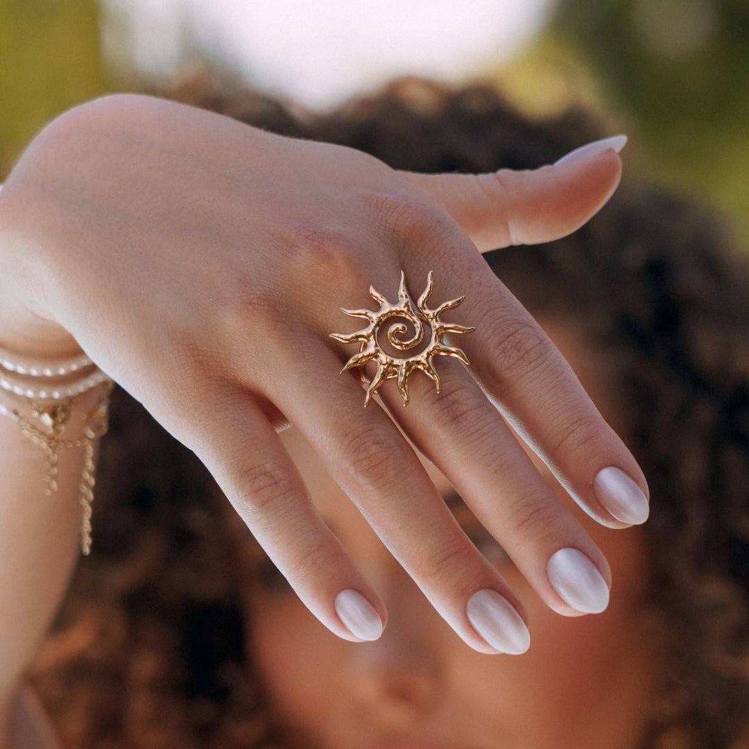 Bague en acier inoxydable doré, motif soleil et spirale, portée sur main féminine.