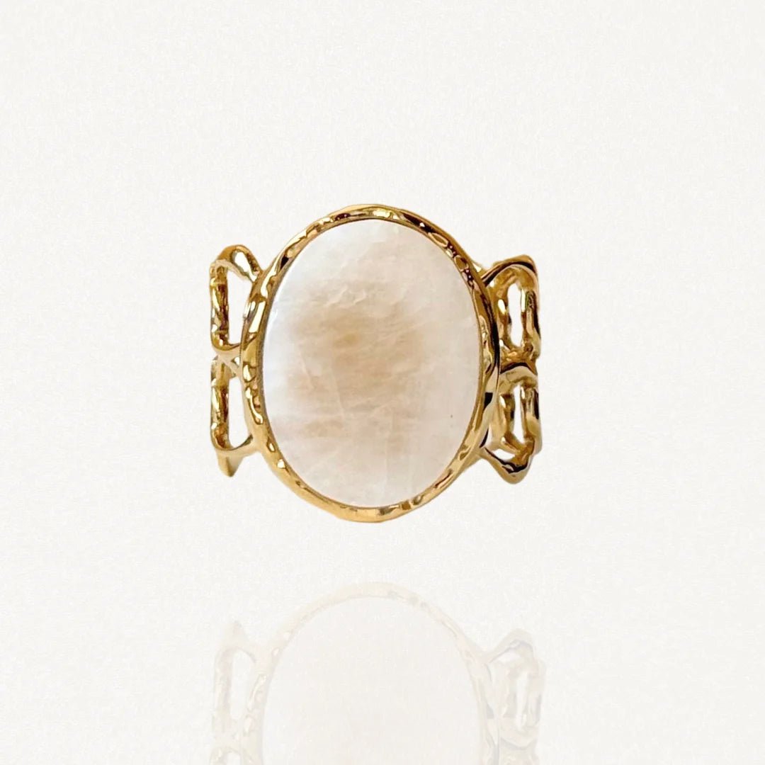 Bague large en acier inoxydable doré ornée d'une grande pierre ronde blanche effet nacre et anneau ajouré texturé sur fond blanc.