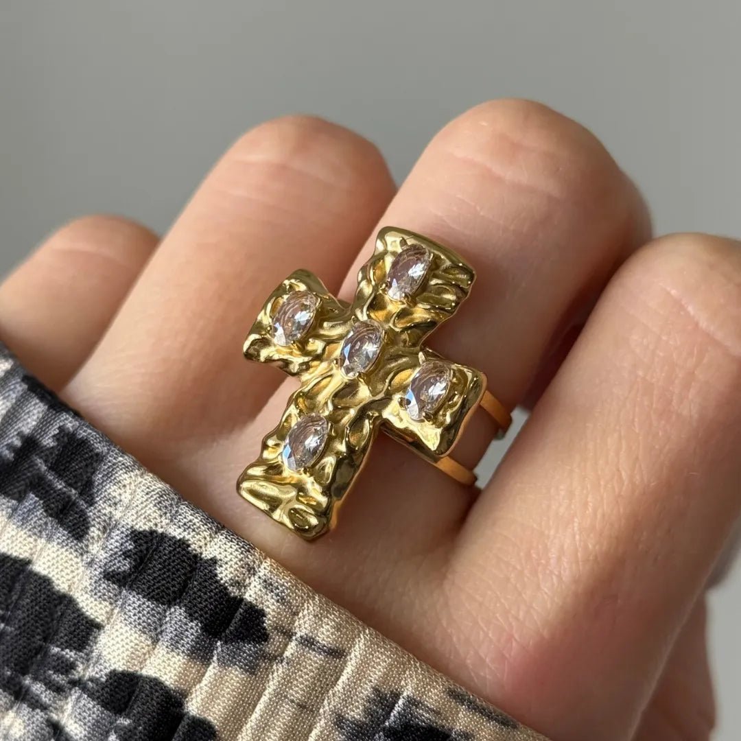 Bague croix dorée martelée, sertie de 5 pierres claires taille rose, portée sur un doigt à côté d'un tissu imprimé animal.