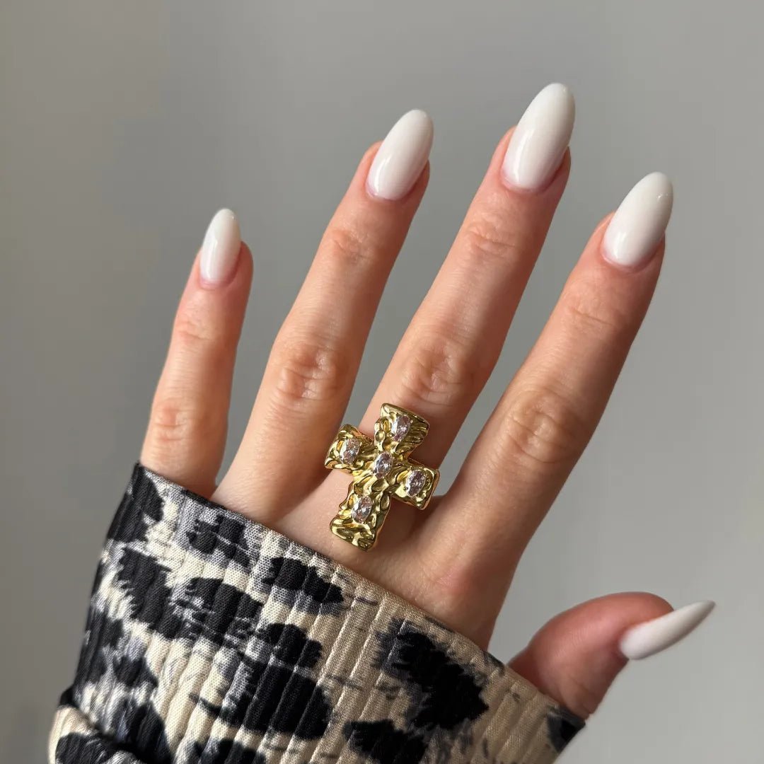 Bague croix or texturée 5 pierres claires sur main aux ongles amande blancs laiteux. Manche imprimé animalier.