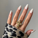 Bague croix or texturée 5 pierres claires sur main aux ongles amande blancs laiteux. Manche imprimé animalier.