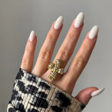 Bague croix or texturée 5 pierres claires sur main aux ongles amande blancs laiteux. Manche imprimé animalier.