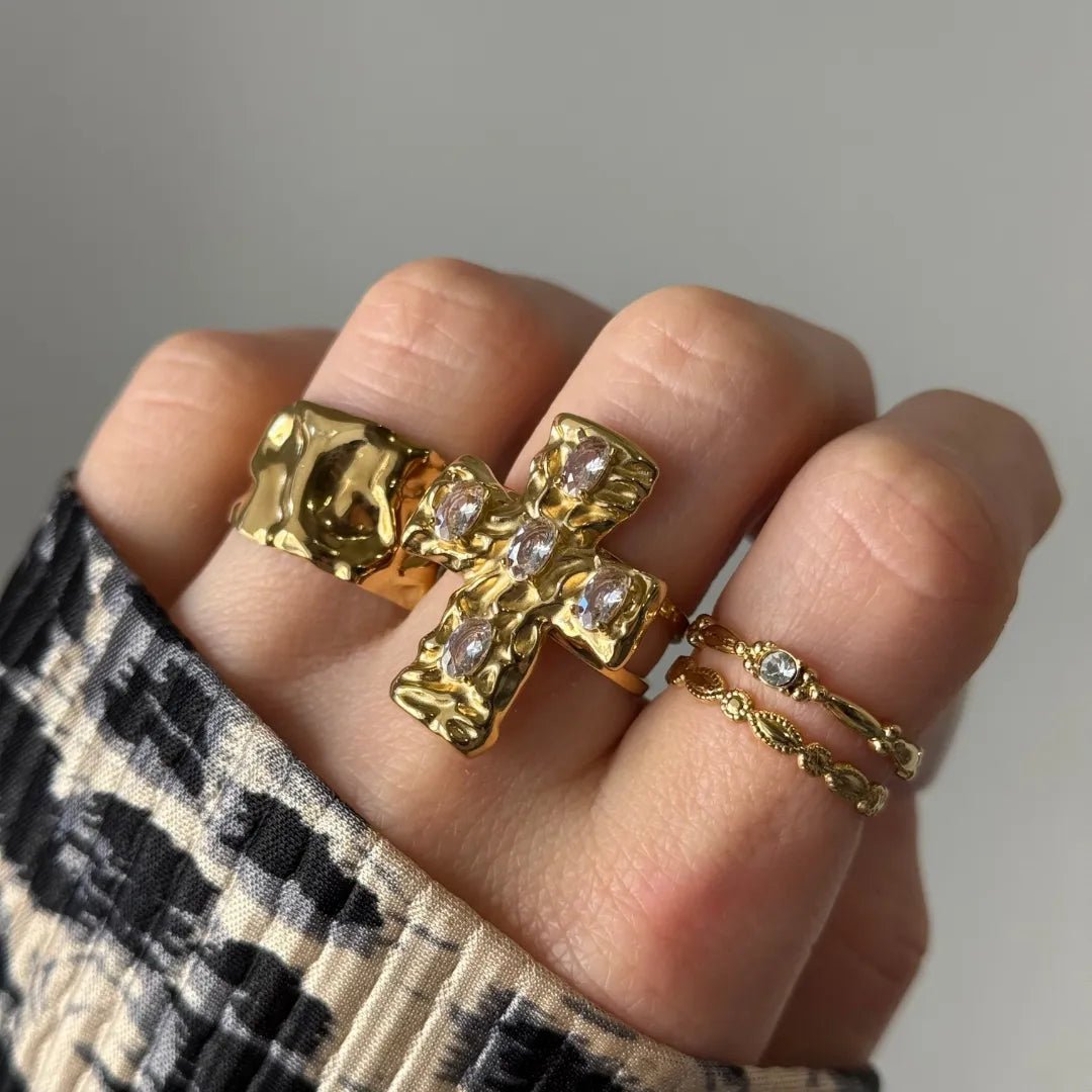 Main de femme portant 4 bagues en or: une martelée large, une croix texturée avec 5 pierres claires, et deux fines, dont une sertie d'une pierre.