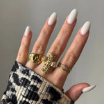 Main aux longs ongles blancs ovales. Trois bagues dorées: chunky texturée, croix sertie de pierres, et double anneau fin. Manche imprimé léopard.