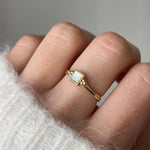 Gros plan bague Melania dorée avec pierre carrée portée au doigt avec un pull blanc douillet.