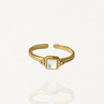 Bague fine ajustable en acier inoxydable doré ornée d'une petite pierre carrée blanche aspect nacre sur fond blanc.