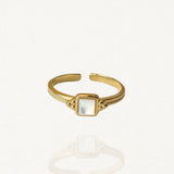 Bague fine ajustable en acier inoxydable doré ornée d'une petite pierre carrée blanche aspect nacre sur fond blanc.