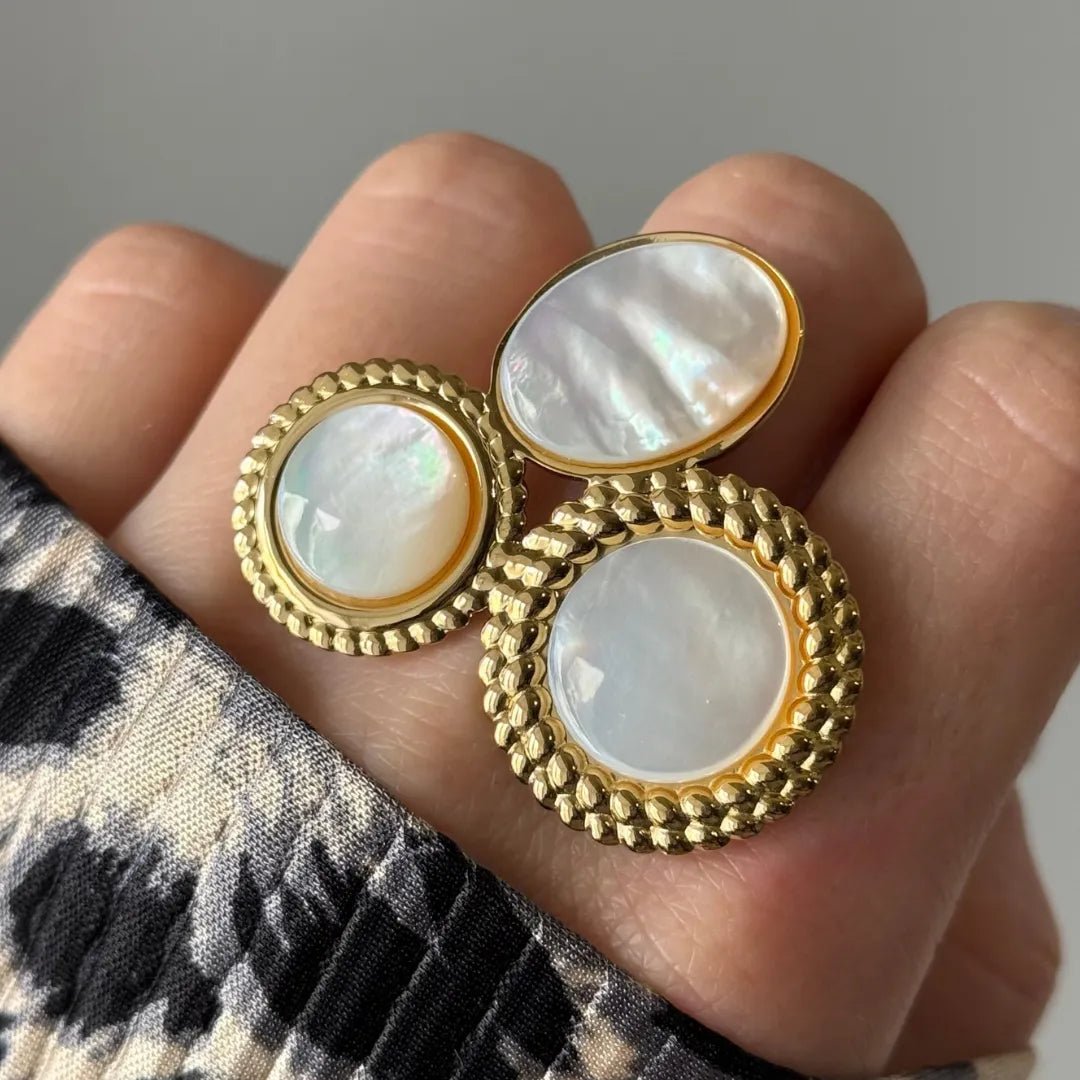 Bijou doré de 3 éléments nacre iridescente: deux ronds, un ovale. Bords texturés torsadés. Sur un doigt, vêtement imprimé.