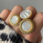 Bijou doré de 3 éléments nacre iridescente: deux ronds, un ovale. Bords texturés torsadés. Sur un doigt, vêtement imprimé.