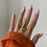 Bague longue Nicola portée en accumulation (stacking) avec d'autres bagues fines dorées sur une main avec manche rouille.