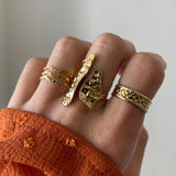 Bague longue Nicola portée en accumulation (stacking) avec d'autres bagues fines dorées sur une main avec manche rouille.