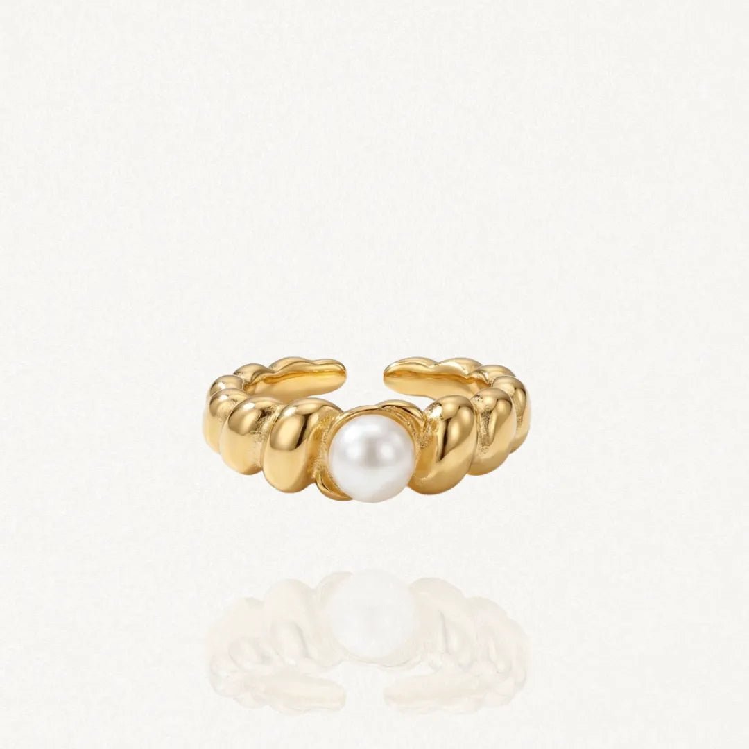Vue rapprochée d'une bague ouverte dorée en acier inoxydable avec un anneau torsadé et une perle blanche ronde au centre. Le bijou est posé sur un fond clair avec un reflet.