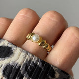 Bague dorée à anneau ondulé et perle blanche centrale portée sur un doigt à côté d'une manche imprimée.