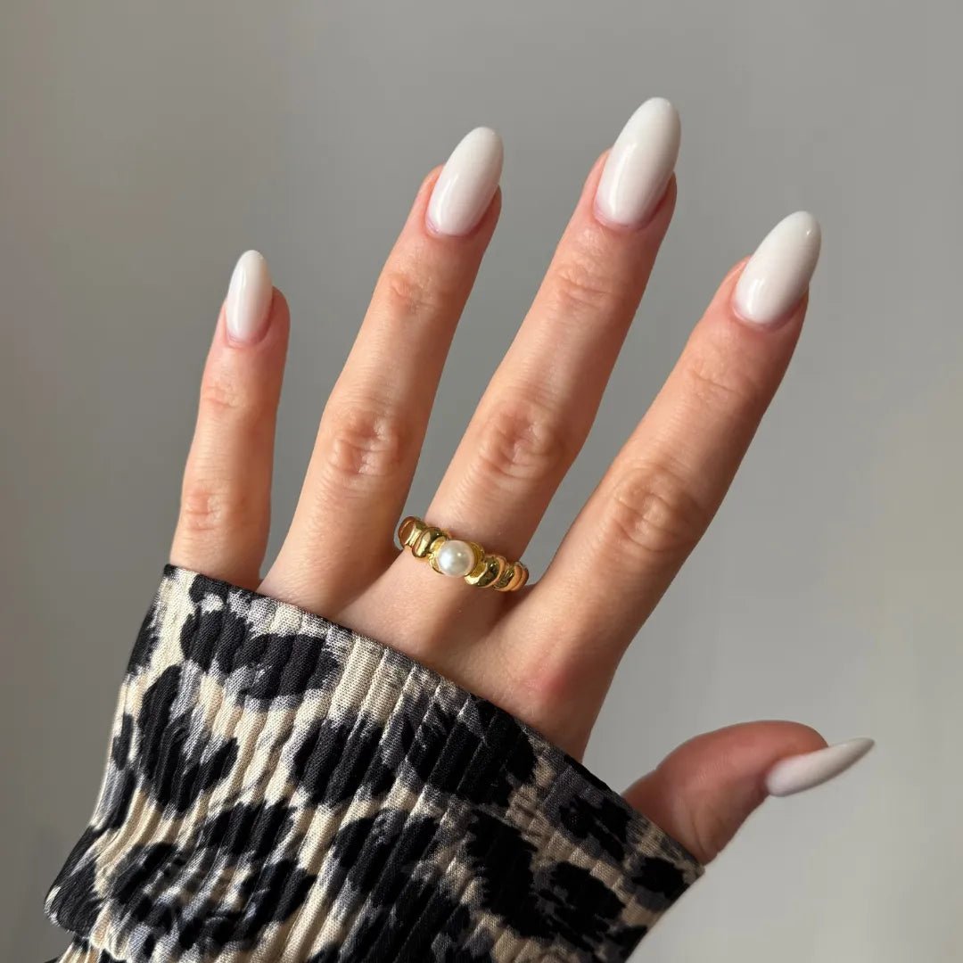 Bague dorée à segments bombés avec perle blanche centrale sur annulaire, main à ongles blancs ovales, manche imprimé léopard.