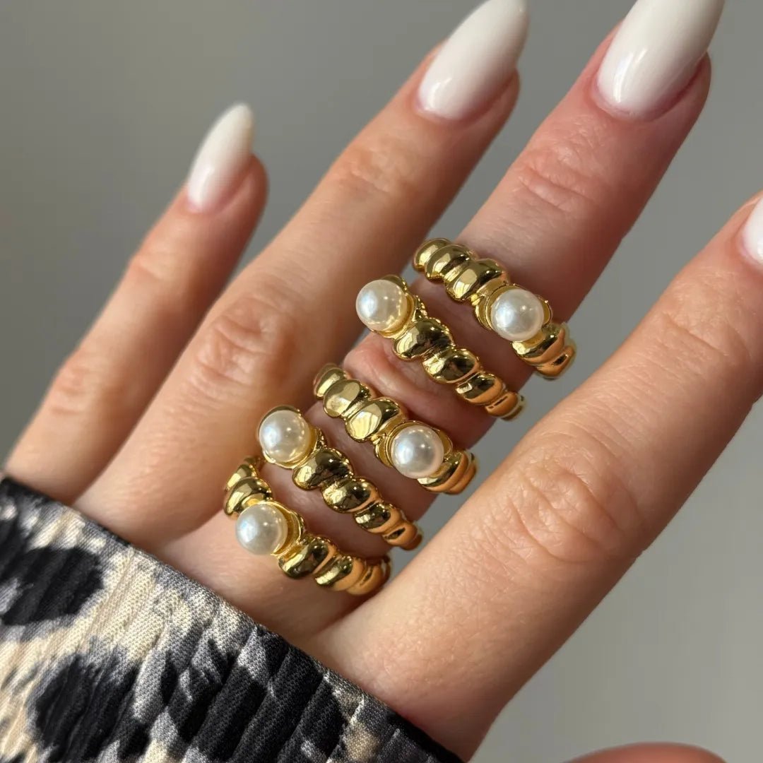Quatre bagues dorées empilées avec perles blanches et anneaux côtelés, portées sur le doigt d'une main aux ongles blancs.