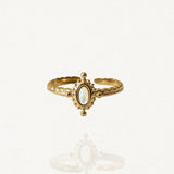 Bague fine ajustable en acier inoxydable doré avec petite pierre ovale blanche et contour perlé style vintage sur fond blanc."