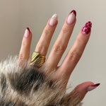 Bague statement dorée au design enveloppant portée sur une main avec une manucure nail art rouge et bordeaux.