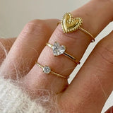 Bague fine Valentina portée en accumulation (stacking) avec d'autres bagues cœurs dorées sur une main.