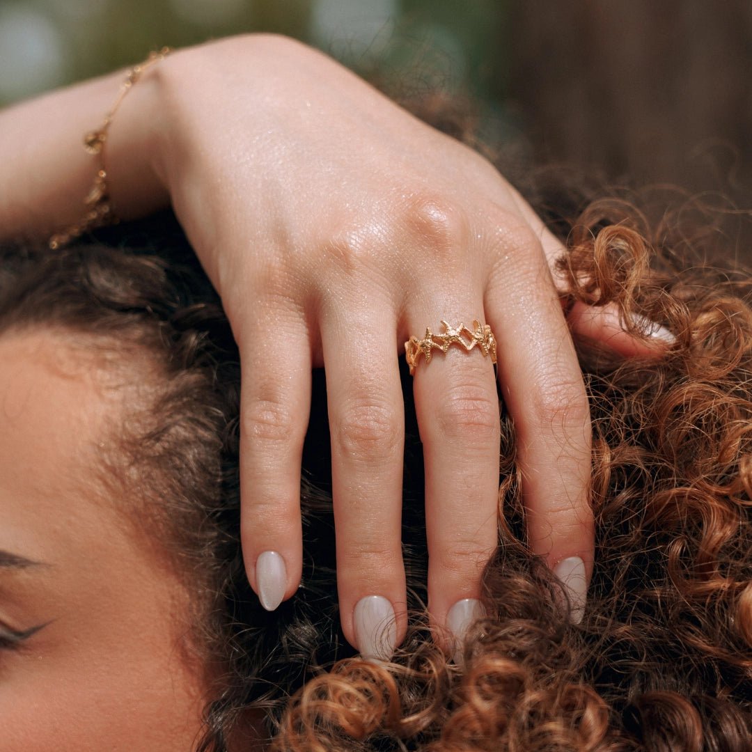 Bague en acier inoxydable dorée, motif ajouré étoilé, portée sur le doigt d'une main avec ongles clairs, sur cheveux bouclés.