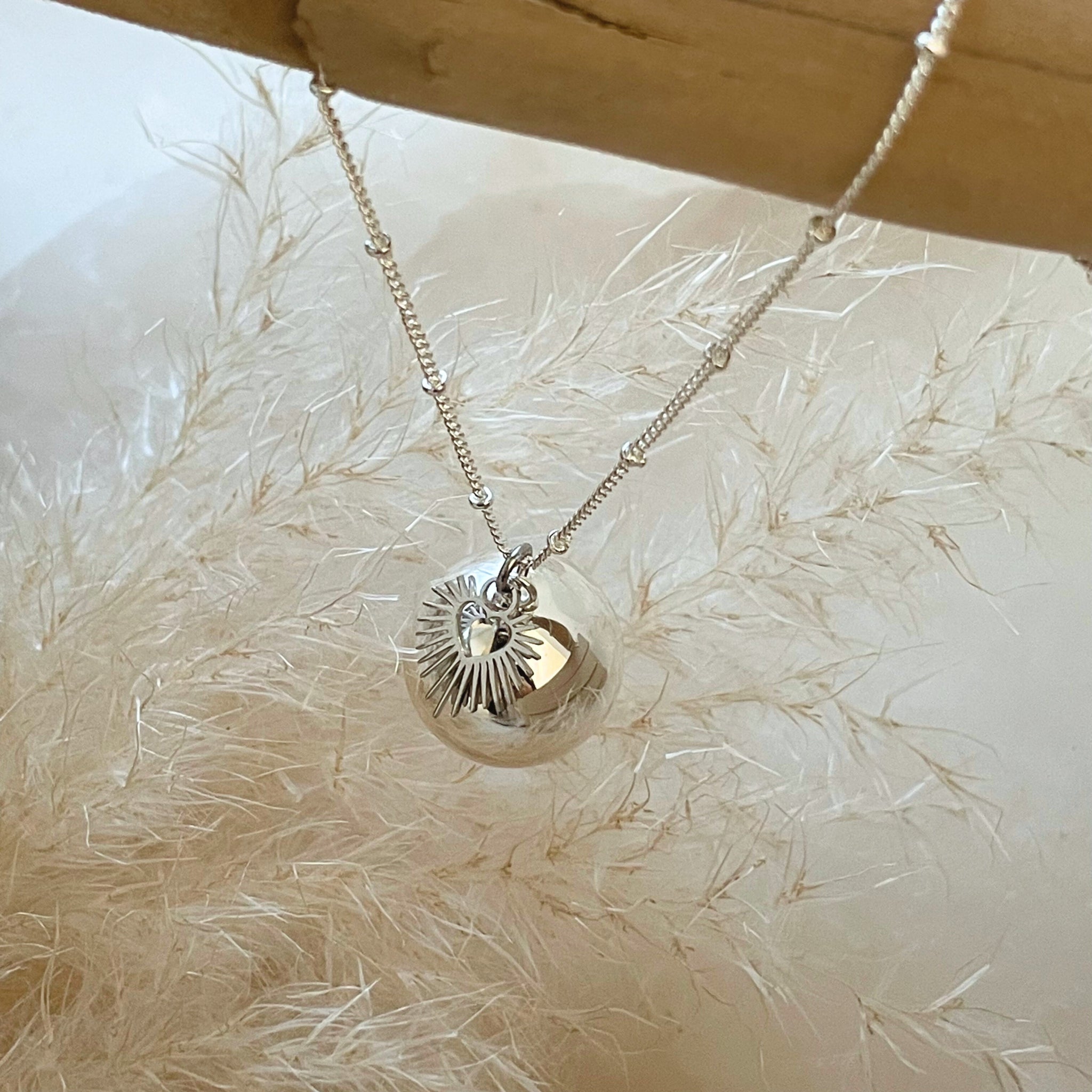 Bola de grossesse en acier inoxydable argenté avec pendentif sonore et breloque cœur sacré rayonnant sur fond blanc.