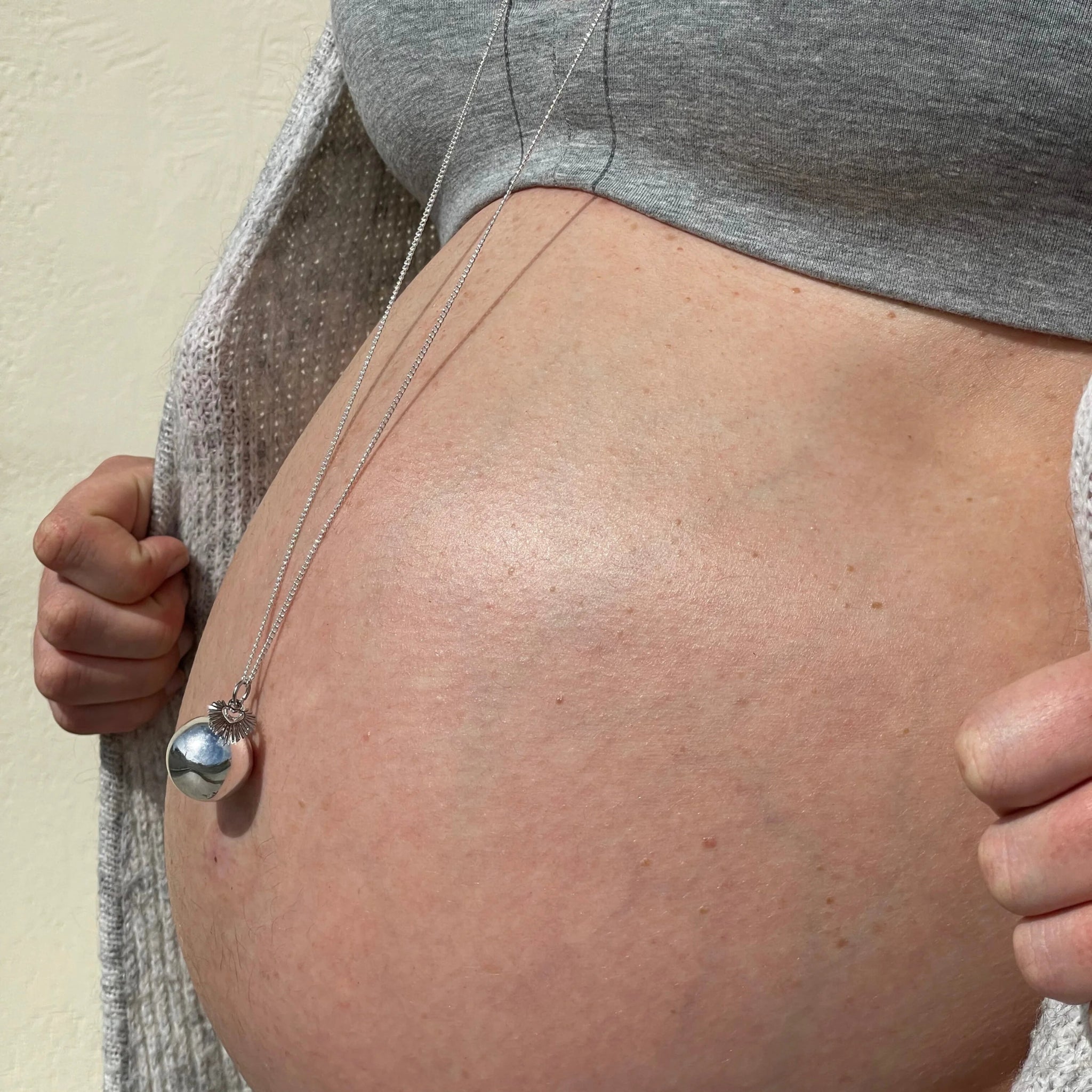 Bola de grossesse argenté porté sur le ventre rond d'une femme enceinte, bijou de maternité apaisant.