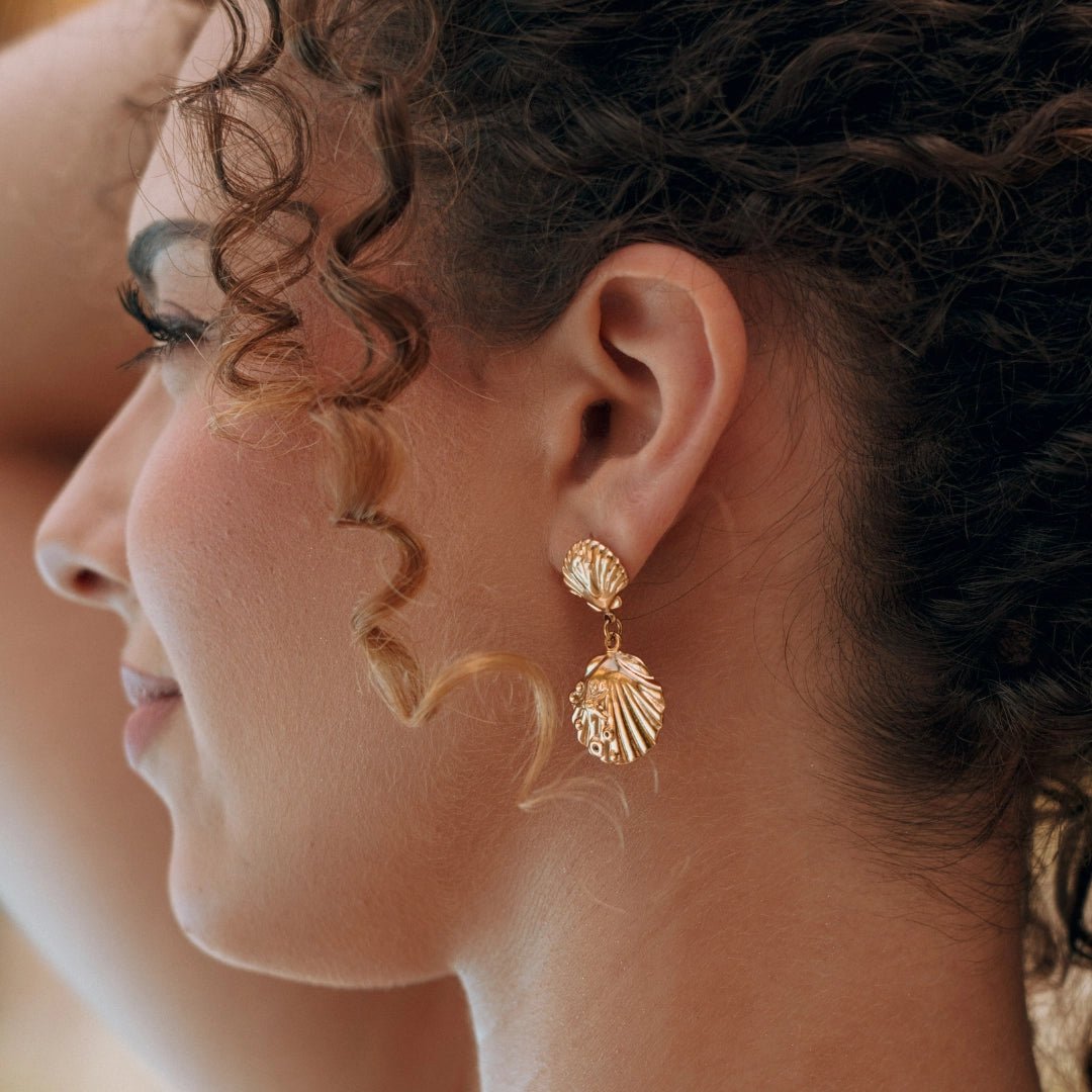 Boucles d'oreilles pendantes en acier inoxydable doré avec motifs coquillages détaillés, portées par une femme.